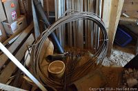 Wide view of a large coil of metal cable stored vertically in a dusty environment, showing the entire length of the coil and parts of surrounding workshop/storage materials.