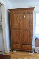 Front view of closed oak armoire showing two paneled doors, crown molding and bottom drawers with wood knobs.