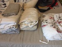 Assorted folded bed linens including three single fitted sheets in cotton and a queen-sized floral duvet cover with two pillow covers, placed on a couch.