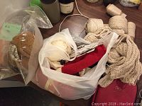 Top view showing plastic bags containing unfinished knitted beige vest, yarn balls and cones along with some spools, surrounded by additional yarn and a bottle