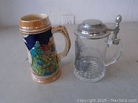Two beer steins on a white surface against a plain wall background. Left: Ceramic stein with colorful medieval village scene. Right: Clear glass stein with hinged pewter lid and etched design.