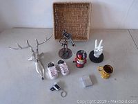 Photo showing all items in the lot including metal deer head, violinist sculpture, baby shoes, helmet figurine, peace sign hand sculpture, glass mug, lighter case, pipe accessory, keychain, and woven basket tray