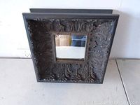 View of square mirror showing the ornate carved floral and scrollwork frame and reflective glass center, placed on a surface with white background.