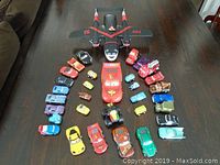 Top view of over 20 die-cast toy cars arranged around a large black toy plane on a wooden table, showing variety of car colors and styles.