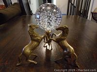 Two brass horse figurines rearing and holding a bubble glass sphere on wooden table.