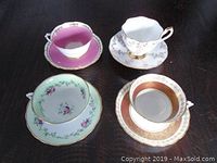 Four different Royal Albert teacups with matching saucers on a dark background, showing colors pink, white with gold, green floral, and gold-brown pattern.