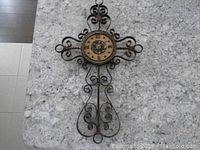 Full view of metal hanging clock on stone countertop showing decorative metal scroll frame and round clock face.