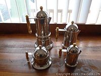 Full view of the four-piece stainless steel set showing all items with gold-tone handles and knobs, on a wooden surface near a window.
