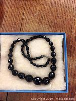 Necklace made of varying sizes of black faceted glass beads, shown coiled on a fabric cushion inside a box.