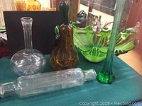 Photo showing six art glass pieces: a pear-shaped amber and green wasp catcher with black stopper, a green ruffled edge bowl, a clear decanter with starburst base, a tall green vase, and two clear bottles of different shapes.