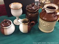 Four pieces of pottery including a two-tone cream and sugar set, a white goblet, a brown coffee holder labeled 'COFFEE', and a large milk pot labeled 'MILK' on a green cloth.