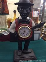 Front view showing the black cast figurine of a man with a round clock inset in his torso, wearing a hat, red vest, white shirt, and holding a guitar.