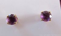 Close view of two round purple amethyst gemstones set in 14K yellow gold prong stud earring settings on white background