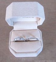 Two silver rings in beige velvet box, one engagement ring with large CZ and one wedding band with smaller CZ stones.