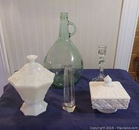Photo showing five items: two milk glass candy dishes with covers, large green glass jug, clear glass candlestick holder, and tall glass bud vase with hexagonal base on a navy blue cloth in front of a white beadboard wall.