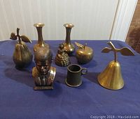 Photo showing all eight brass items on a blue surface against a wall: two pear-shaped bells, two narrow-necked vases, J.F. Kennedy bust, woman figurine, brass apple, and a small textured cup.