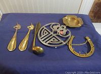 Full lot on blue background showing all six brass items including two unicorn-handled shoe horns, ladle, trivet, ashtray, and horseshoe key holder.