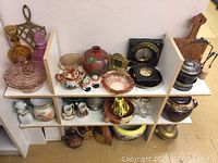 Photo showing assorted collectibles arranged on white shelving including pottery, glassware, figurines, metal and wood items.