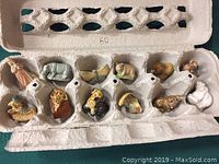 View of all 12 small ceramic tea figurines arranged inside an egg carton container, showing variety of shapes and colors.