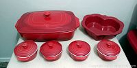 Photo shows six red ceramic casserole dishes: one large rectangular with lid, one heart-shaped with lid, and four small round covered dishes, all arranged on a table.