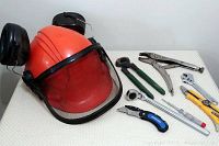 Full view of safety helmet with ear protection and mesh face shield, laid flat alongside pliers, wrench, utility knife, measuring tape, and pocket knife.
