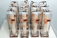 Photo showing 12 tall slender white candles with orange floral patterns in clear plastic cylindrical containers with brown bases and black string closures, arranged in rows.