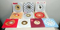 Ten vintage 45 RPM records with various record company paper sleeves arranged in two rows on white surface.