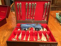 Overall view of the 1847 Rogers Bros flatware set displayed in open wooden chest with red velvet lining, showing knives in lid panel and forks, spoons in lower compartments along with fabric silverware roll.