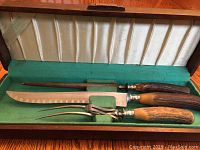 Photo showing two complete carving sets with knives, forks, and sharpening steels in a wooden box with green and white fabric lining.