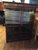 Front view of top section of two-piece cabinet with double glass paneled doors wrapped in clear plastic film.