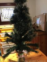 Full view of a 48 inch tall artificial fibre optic Christmas tree in a gold and white decorative base, standing indoors near a window and staircase.