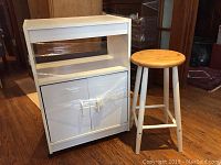 Front view of white rolling kitchen cart with wooden stool beside it on wood floor.