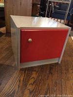 Solid wooden end table cabinet with light wood finish and single red door with metal knob visible from front and angled side view.