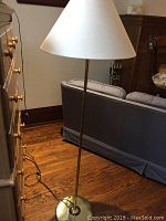 Photo of tall brass floor lamp with round base and beige tapered shade with a dent.