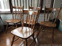 Four solid oak wooden chairs arranged together showing the overall design and condition. Classic wooden spindle back chairs with a warm finish.