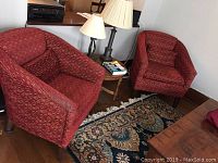 Two red upholstered club chairs placed side by side in a room with wooden floor, a small table with lamps and books between them.