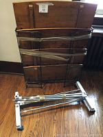 Metal folding shoe rack with wood slat shelves laying against wall, taped together with parts of silver metal rolling clothing rack on the floor in front.