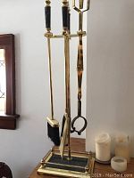 Photo of fireplace tools set showing broom, poker, and tongs with marble handles hanging on the brass and marble stand, placed near a window and candles.