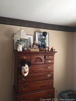 Wide shot showing the chest with two lamps, two framed prints, a carved mask hanging on the side, small figurines, a model sailboat and a rickshaw model on top of the chest of drawers.