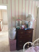 Full view of room corner showing hardwood dresser with wicker hampers beside it, large glass jar on top of dresser, floral lamp, and smaller decor items.