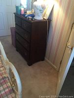 Full view of dark wooden dresser standing on carpeted floor near striped wallpaper wall, showing front and side with visible wear and brass drawer handles