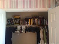 Wide shot showing closet shelf with a variety of books, encyclopedias, and magazines arranged along with hanging clothes below.