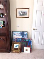 Framed artwork and mirror grouped near a white door and wooden cabinet. Includes gold ornate mirror and multiple framed paintings leaning on the floor.