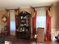 Full view of one parlor window with rust-colored drapes, beige and red valance swags with tassels, sheer white curtains, antique wooden furniture in front.