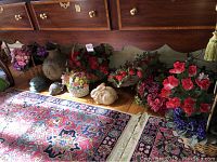Photo showing multiple artificial flower arrangements with red and purple flowers in baskets, cement bunny statue, and clay pot under wooden furniture.