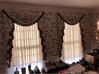 Two dining room windows with window treatments including floral patterned curtains, matching swag valences with tassel trim, and sheer white panels.