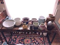 Collection of 11 pottery items including bowls, vases, souvenir pitcher and wine chiller arranged on a rustic table with floral carpet below.