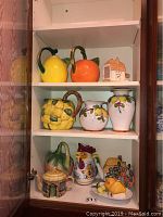 Shelf with brightly colored ceramic pitchers and decorative items including lemon and orange-shaped pitchers, vases with lemon motifs, and whimsical ceramic containers shaped like a rooster and houses.