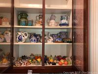 Full view of wooden display cabinet with several shelves holding ceramic and stoneware pitchers, salt and pepper shakers, and miniature figurines, showing the variety of items in the lot.