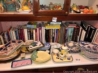 Books arranged on shelves with various ceramic dishes in front. Titles visible from multiple genres including history and fiction.